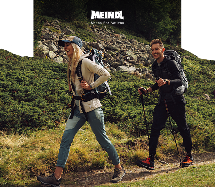 Two hikers wearing backpacks and trekking poles walking on a rocky mountain trail; close-up of Meindl hiking shoes and outdoor gear.