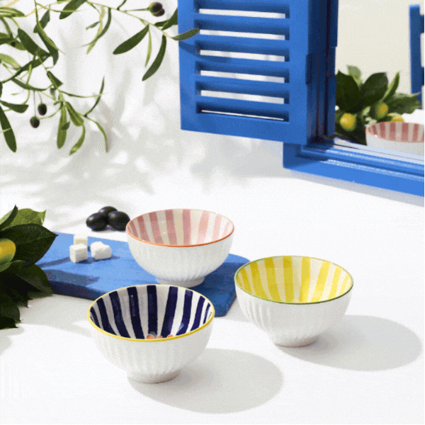 Ceramic bowls and plates with striped sunburst patterns and multilingual phrases beside a blue shutter and olive sprigs
