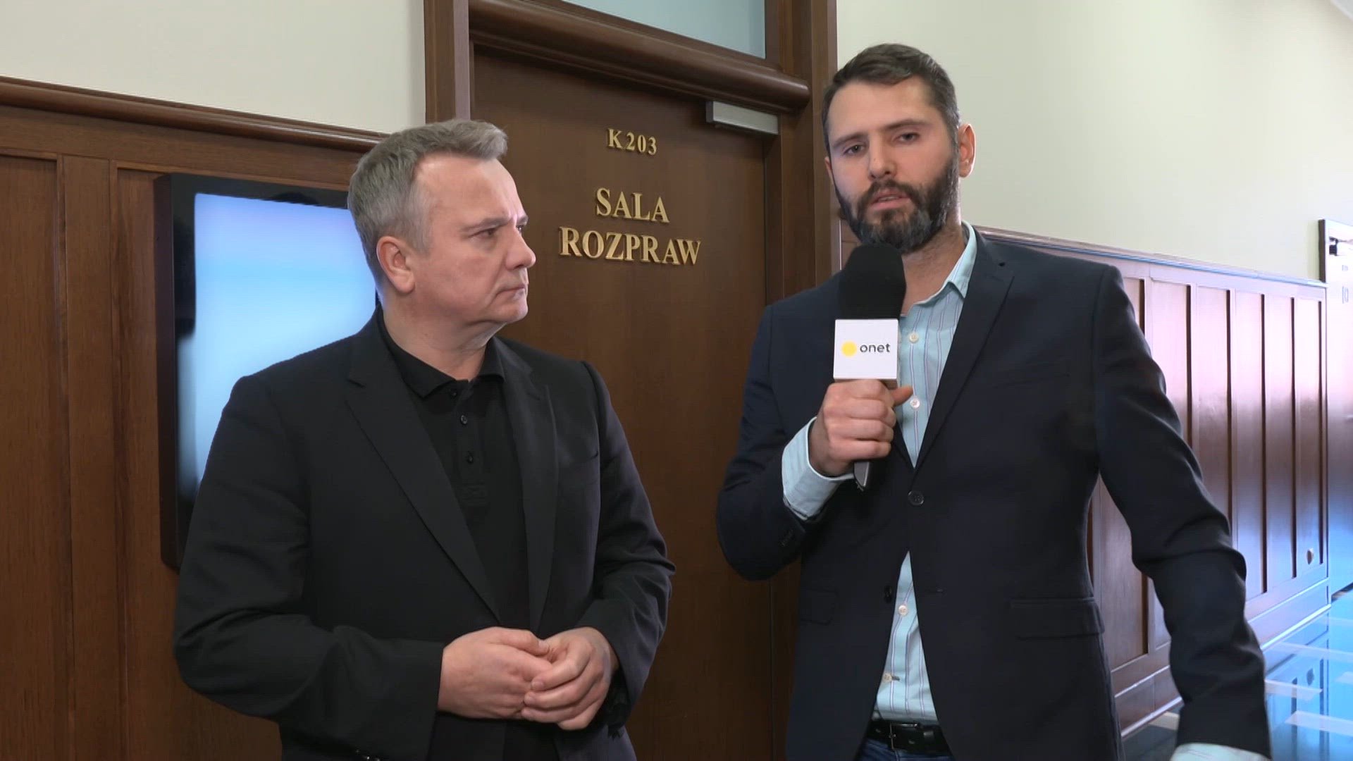 Dwóch mężczyzn w garniturach przed drzwiami sali rozpraw, jeden trzyma mikrofon z logo Onet podczas relacji prasowej