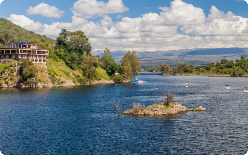 Lakeside building on a green hillside, small rocky islet and boats on a blue lake with distant mountains under a cloudy sky