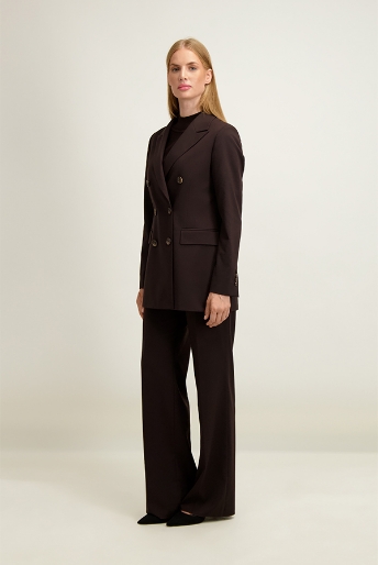 Full-length shot of a blonde woman standing in a dark brown double-breasted blazer and wide-leg trousers with a black top on a plain background