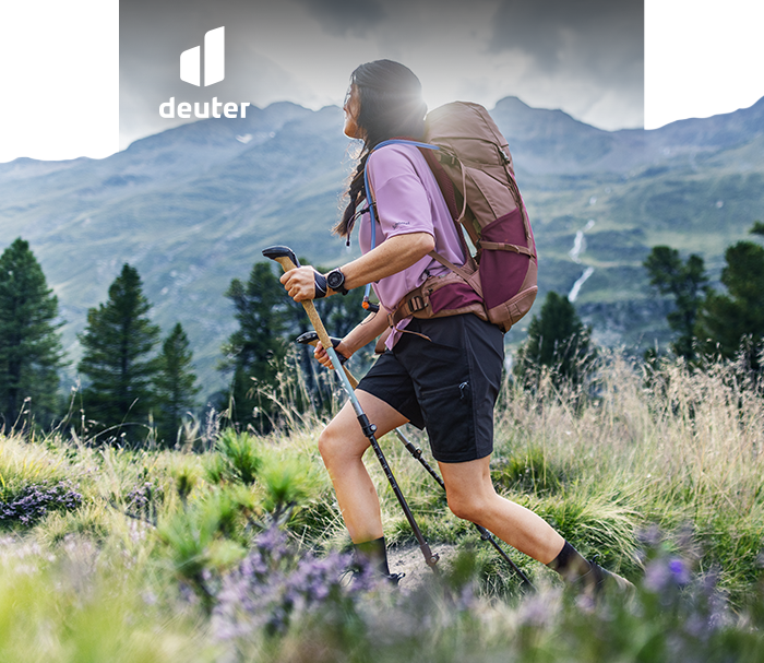 Hiker with large Deuter backpack using trekking poles on alpine trail, wildflowers in foreground and mountains in background