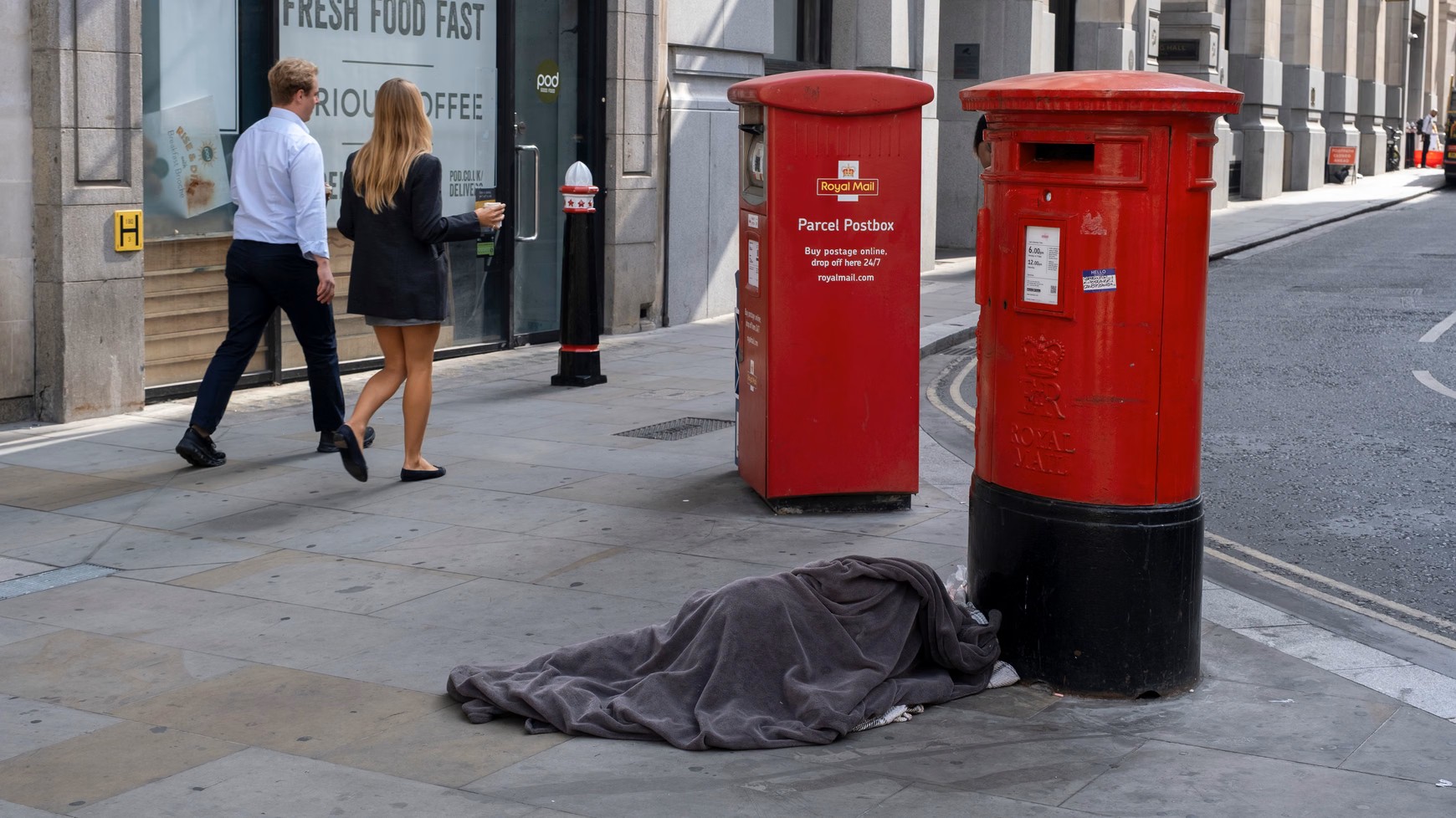 Osoba bezdomna śpi pod kocem obok czerwonych skrzynek pocztowych Royal Mail na chodniku w Londynie, mijana przez przechodniów
