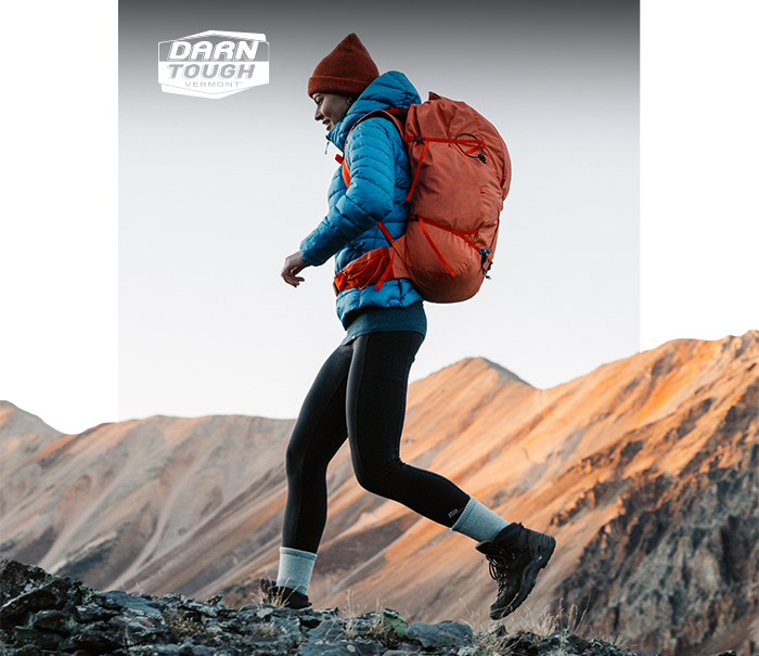 Hiker in blue jacket, red backpack and Darn Tough socks trekking across a rocky ridge with sunlit mountain slopes