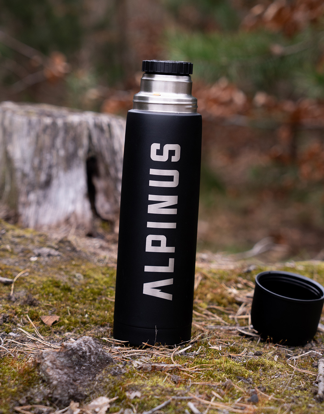Black Alpinus insulated flask standing on mossy forest ground beside its detached cup, stainless-steel rim visible