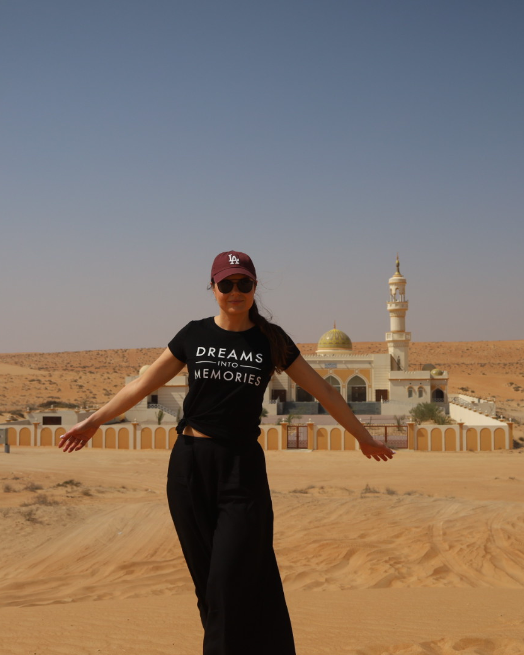 Woman wearing 'Dreams into Memories' T-shirt and baseball cap standing with arms outstretched in desert in front of a mosque