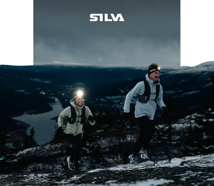 Two trail runners wearing headlamps on a snowy hillside at dusk, lake below and SILVA logo above