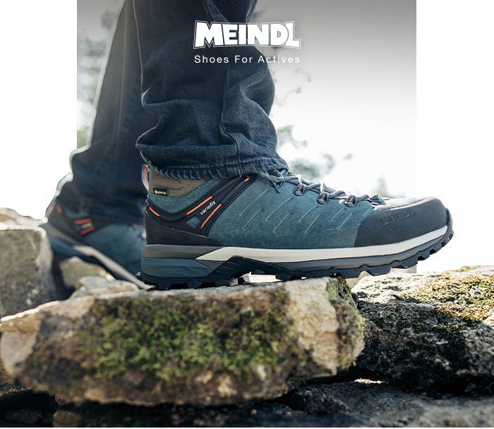 Close-up of blue Meindl hiking boot with Gore‑Tex label on a rocky, mossy trail, person wearing jeans