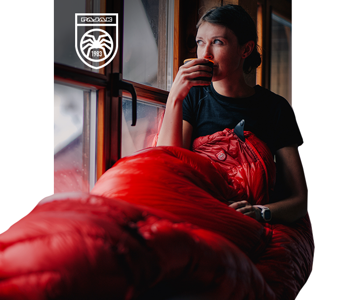 Woman wrapped in a red sleeping bag beside a window, sipping a hot drink — cosy outdoor sleeping-bag lifestyle with brand logo