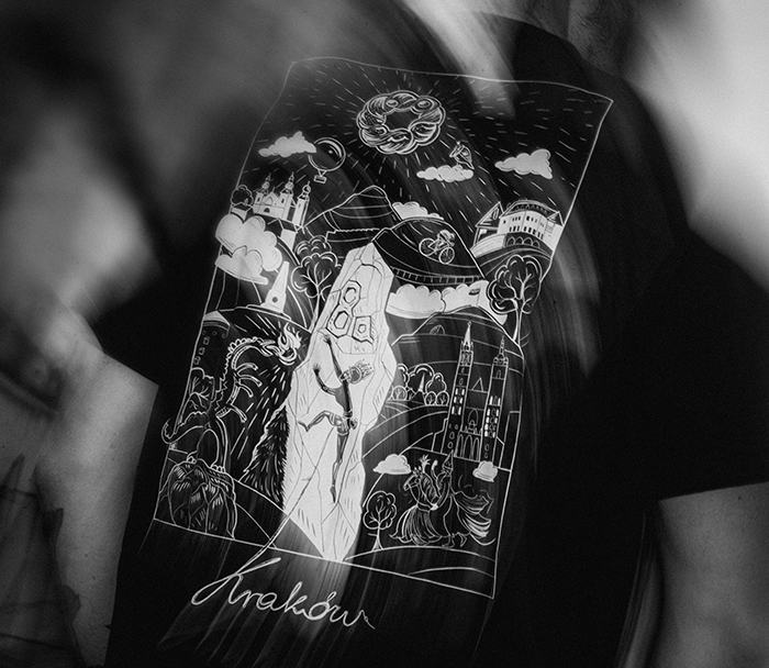 Black T‑shirt with white Kraków illustration: climber on a mountain amid city landmarks and clouds, photographed on a wearer