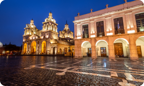 Illuminated colonial cathedral and arched municipal building by wet cobblestone plaza at dusk