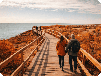 Couple walking along a wooden coastal boardwalk amid autumn foliage with sea view under a cloudy sky