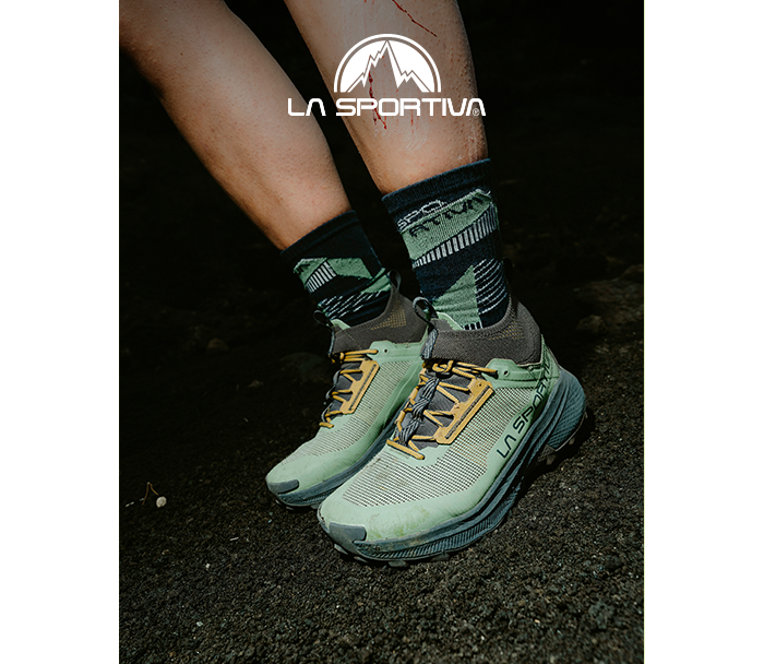 Close-up of green La Sportiva trail running shoes on feet with patterned socks standing on dark, muddy trail