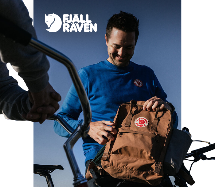 Smiling man on a bicycle adjusting a brown Fjällräven Kånken backpack with visible logo against a clear blue sky