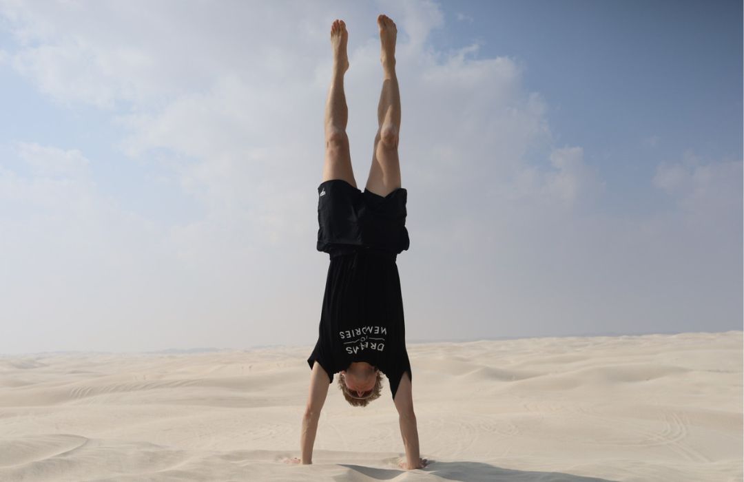 Person performing a handstand in black shorts and t-shirt on expansive white sand dunes under a blue sky