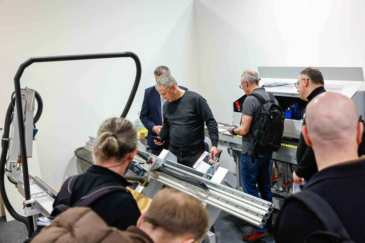Group of people inspecting an industrial paper-folding machine at a trade show, man holding brochure and smartphone
