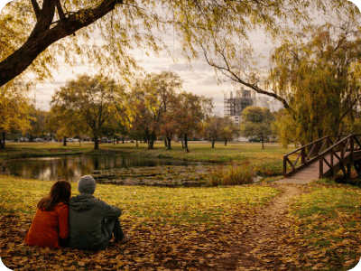 Couple sitting on leafy bank beside a pond in an autumn park near a wooden footbridge with city buildings beyond