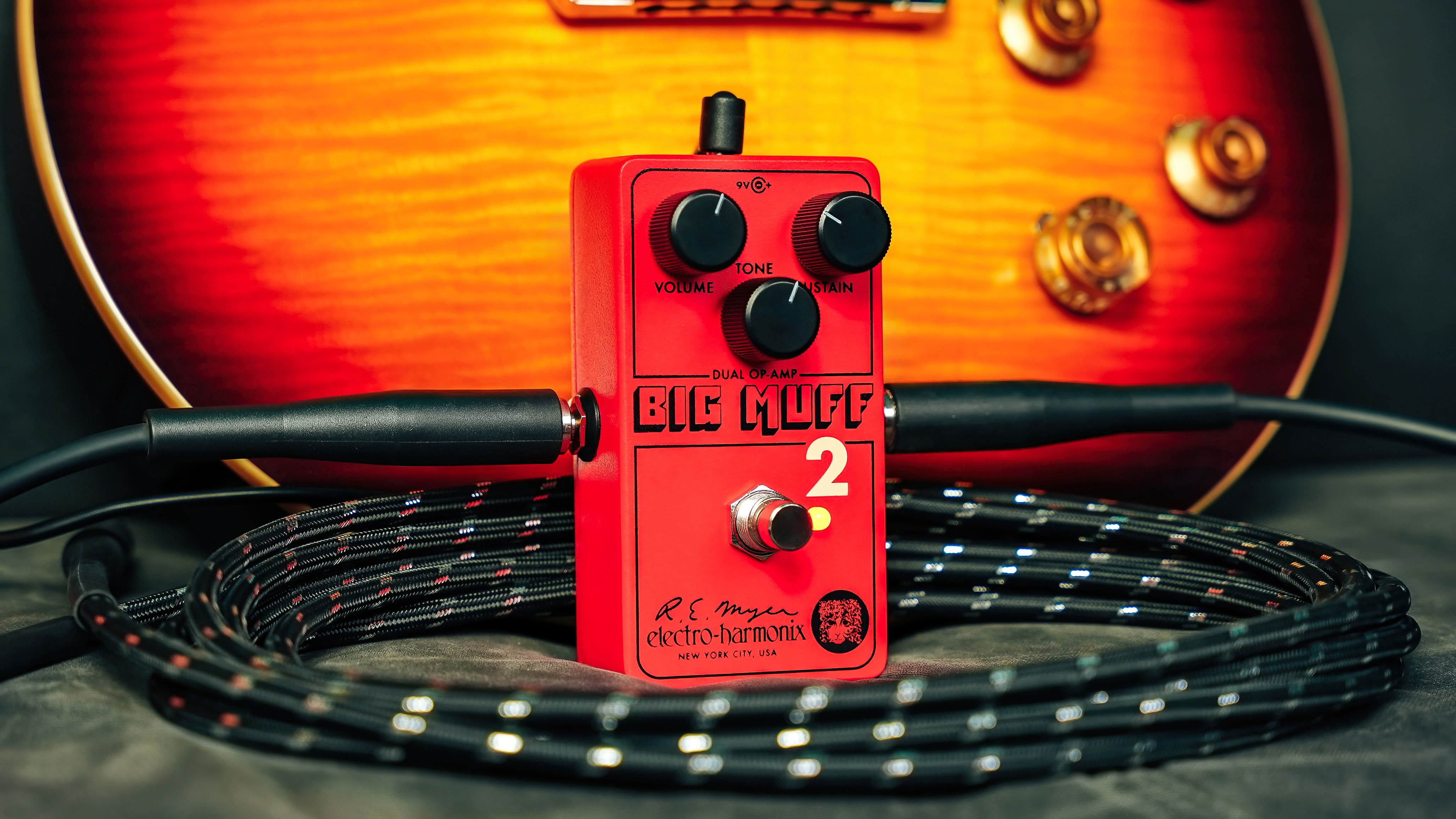 Red Big Muff 2 guitar distortion pedal with knobs and footswitch, coiled instrument cable and sunburst electric guitar in the background